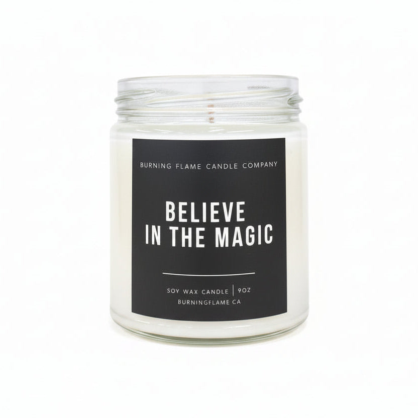 A 9oz soy wax candle in a clear glass jar with a black lid, featuring a black label with white text that reads 'BELIEVE IN THE MAGIC' by Burning Flame Candle Company.