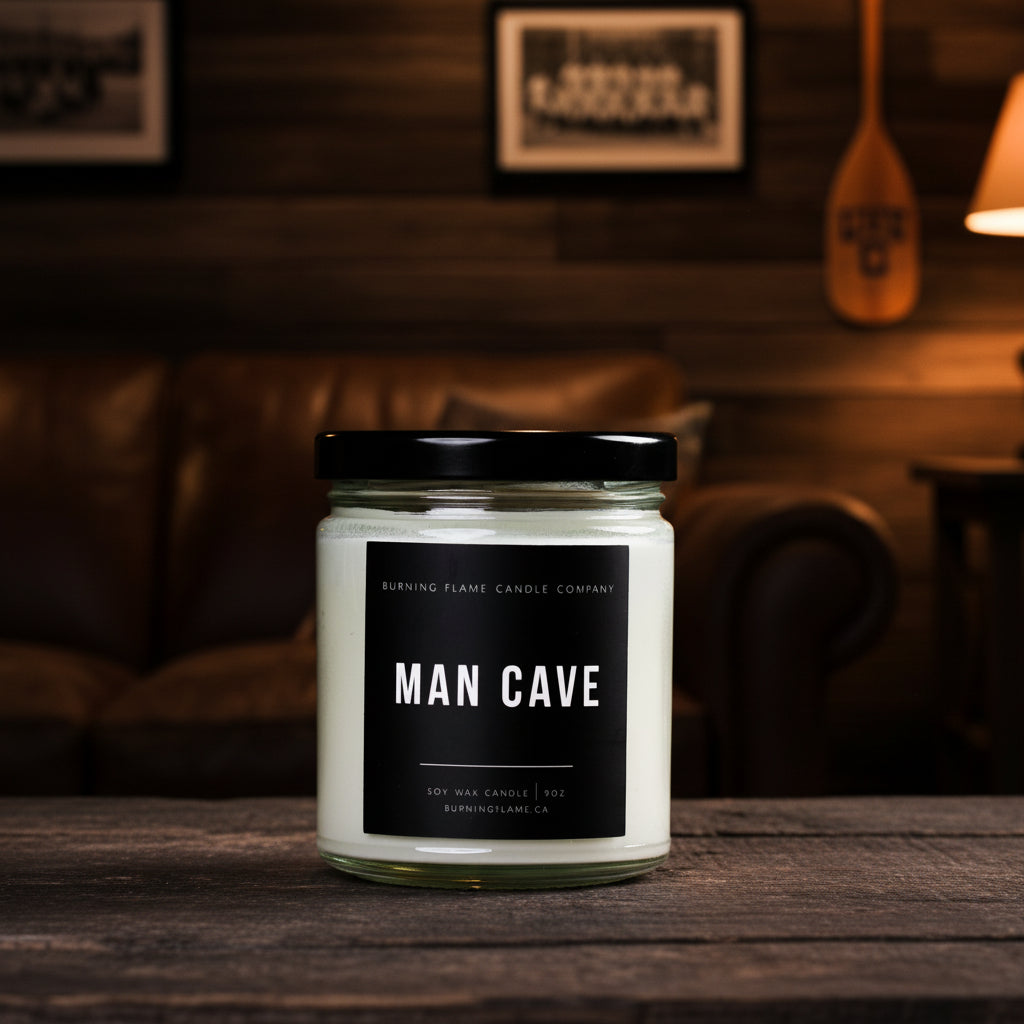 A jar candle labeled 'MAN CAVE' by Burning Flame Candle Company sits on a dark wooden table in a cozy, dimly lit living room with a leather couch and framed wall art in the background.