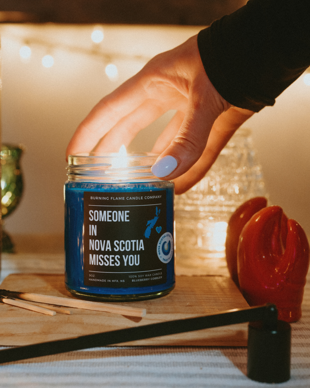 Hand holding the Someoe in Nova Scotia Misses you candle jar, set against a warm, indoor background.