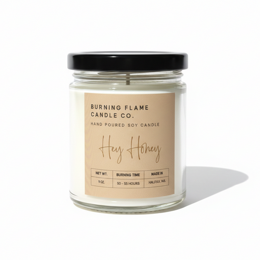 A glass jar candle with a black lid and a beige label reading 'BURNING FLAME CANDLE CO. Hey Honey' on a clean white background.